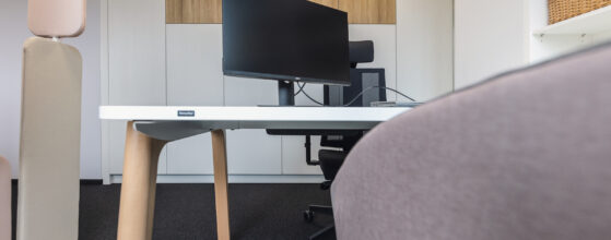 Nowoczesna przestrzeń biurowa z białym biurkiem, monitorem i ergonomicznym krzesłem. Czysty design z drewnianymi akcentami.